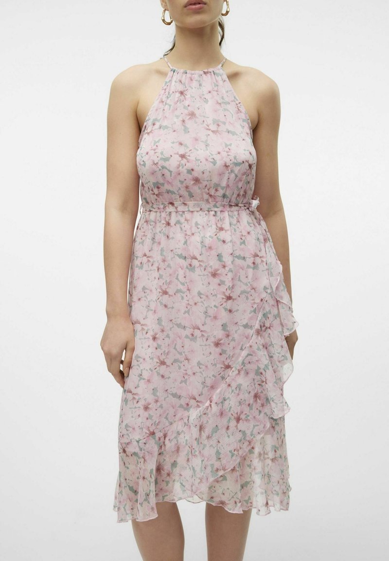 Femme portant une robe sans manches à bretelles halter rose clair à motif floral, avec des détails volantés et des boucles d'oreilles créoles dorées sur un fond blanc uni.