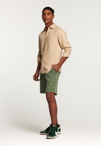 Camicia beige a maniche lunghe, pantaloni corti verdi con motivo a diamante, scarpe da ginnastica nere e verdi con dettagli bianchi, in posa in piedi.