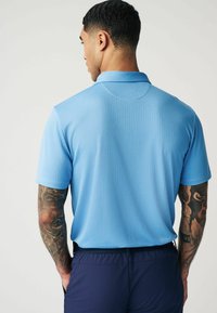 Ein hellblaues Poloshirt mit einem strukturierten Muster, kurzen Ärmeln und einem klassischen Kragen, kombiniert mit navyblauen Hosen. Sichtbare Tattoos am Arm.
