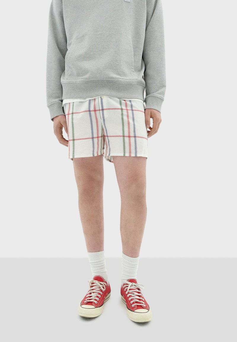 Grijze sweatshirt, geblokte short in wit met rode, groene en blauwe lijnen, witte sneakersokken en rode canvas sneakers met witte zolen.
