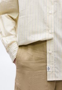 Personne portant une chemise à rayures beige et marine légèrement rentrée dans un pantalon marron clair texturé, une main dans la poche.