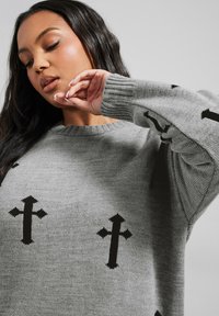 Grå stickad tröja med rundad halsringning, lösa ärmar och svarta korsmönster överallt. Mjuk textur och avslappnad passform.