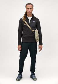 Pullover in pile nero con zip a un quarto, tessuto testurizzato; borsa a spalla verde; pantaloni neri; scarpe a fantasia grigie e verdi.