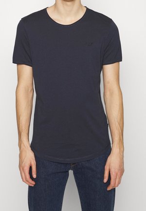 Homme portant un t-shirt uni bleu marine foncé à manches courtes et un jean bleu foncé, debout devant un fond clair.