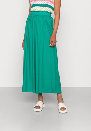 Femme portant une jupe bleu sarcelle taille haute jusqu’aux chevilles, un haut sans manches rayé multicolore et des sandales blanches à bout ouvert, debout sur un sol gris.