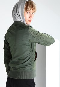 Zelená bomber bunda s rebrovaným lemom a manžetami, ktorá má zipsovú vrecko a elegantnú, hladkú textúru. Kapucňový dizajn zvyšuje všestrannosť.