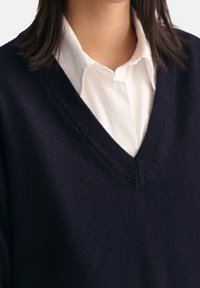 Marineblauer gestrickter V-Ausschnitt-Pullover aus weichem Material, übereinander getragen mit einem weißen Hemd mit Kragen, mit gerippten Bündchen und Saum.