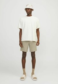 Brīvi pieguļošs balts T-krekls, bēšas teksturētas šortus, bēšas zeķes un baltas čības; nēsā viegli rakstainu cepuri ar platu malu.