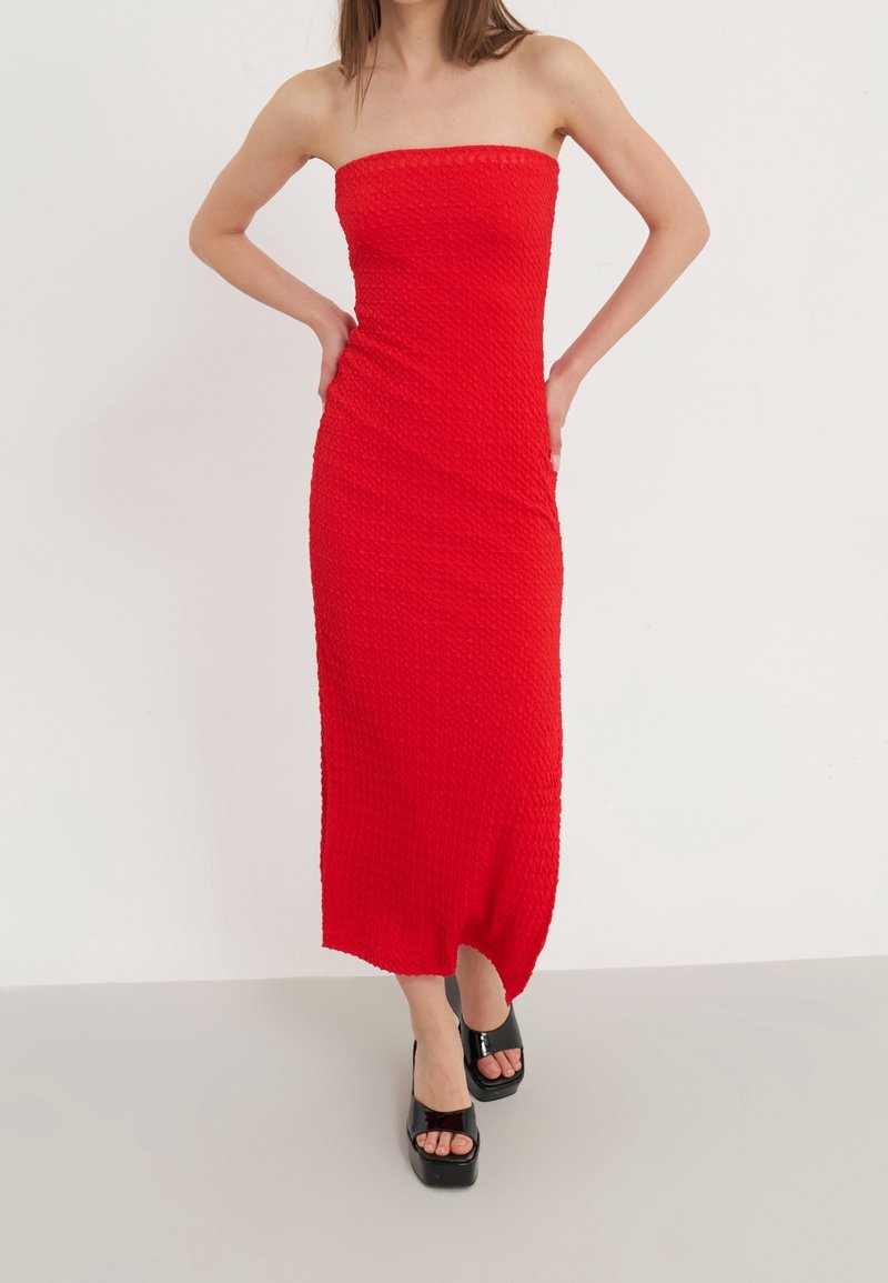 Femme portant une robe midi rouge texturée sans bretelles, associée à des talons à plateforme noirs ouverts à l’avant, debout devant un fond uni.
