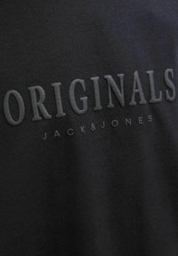 Černá mikina s texturovaným, matně černým písmem, které zní "ORIGINALS" a "JACK & JONES" pod ním v decentním, minimalistickém designu.