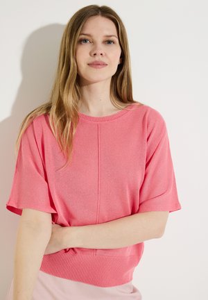 Jeune femme aux longs cheveux blonds portant un pull rose ample à manches courtes, debout avec un bras croisé devant un fond blanc uni.