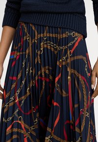 Ráncolt tengerészkék szoknya piros, barna és arany lánc- és övmintákkal, sötétkék, texturált kötött pulóverrel párosítva.