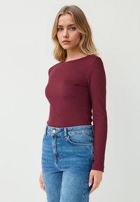 Langärmliges, geripptes Oberteil in tiefburgunderrot, kombiniert mit hoch taillierten hellblauen Jeans. Der Stoff wirkt glatt und hat eine figurbetonte Passform.