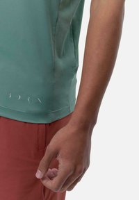 Zaļa sporta krekls ar teksturētu sānu paneli, ar atstarojošu zīmolu apakšmalā. Īsām piedurknēm un kopā ar rūsu krāsas biksēm.