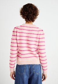 Persoon met krullend haar, gekleed in een gestreepte trui in roze en paars en blauwe spijkerbroek, met de rug naar voren tegen een effen achtergrond.