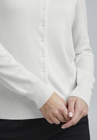 Cardigan bianco realizzato in tessuto morbido, con chiusura a bottoni, polsini e vita a coste. Tessuto liscio senza motivi.