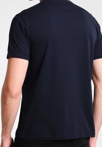 Muž v jednoduchém tmavě modrém tričku s krátkým rukávem, zobrazený zezadu, na jednobarevném světlém pozadí.