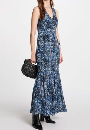 Frau in einem ärmellosen blau-schwarz gemusterten Maxikleid, die eine schwarze, strukturierte Handtasche hält, schwarze Stiefel mit Absatz trägt und eine silberne Halskette trägt.
