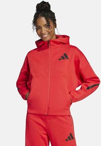 Rote Reißverschlussjacke mit Kapuze, schwarzen Akzentstreifen an den Ärmeln und Logodetail auf der Brust. Aus strukturiertem Stoff gefertigt, lässige Passform.