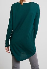 Langärmliges, dunkeltealfarbenes Oberteil mit weitem Ausschnitt und abgerundetem Saum. Weicher, dehnbarer Stoff mit glatter Oberfläche und überschnittenen Schulternähten.