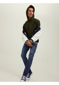 Felpa verde con cappuccio e cordini, giacca varsity blu e bianca, jeans blu strappati e sneakers navy. Focalizzati su uno stile streetwear casual.