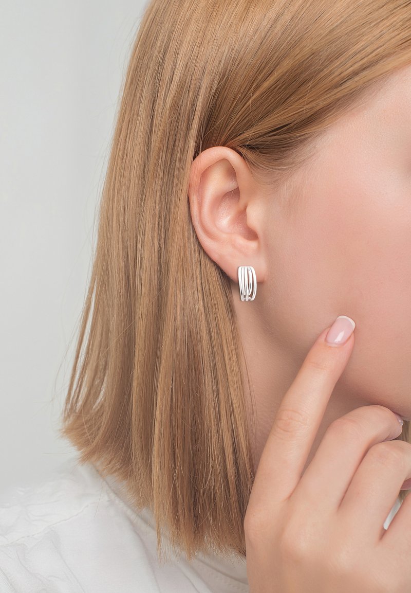Orecchini a cerchio argentati con un design curvato a strati. Superficie liscia e lucida; mostrati su un modello con capelli lisci e castano chiaro.