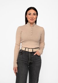Frau mit glattem dunklem Haar trägt einen beige gerippten Pullover mit Knöpfen, schwarze hochtaillierte Jeans und einen beigen Gürtel, lächelt und hat eine Hand in der Tasche.