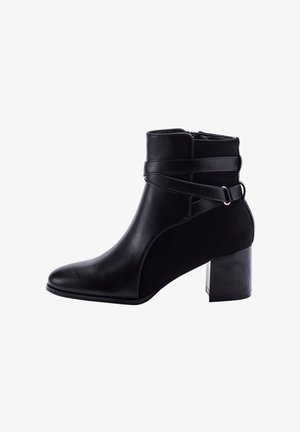 Bottine noire en simili cuir texturé. Présente un bout pointu, un talon épais et un détail de sangle croisée avec des accessoires en métal.