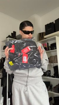 Schwarze Kleidung in einer transparenten Plastiktüte, mit mehreren Preisschildern. Das Model trägt einen grauen Reißverschluss-Hoodie und dunkle Sonnenbrillen.
