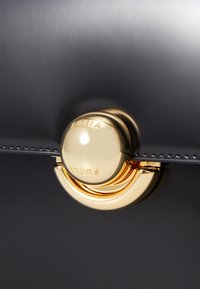 Borsa a mano in pelle nera con finitura lucida, caratterizzata da una chiusura circolare dorata impreziosita dal logo "Furla". Design elegante e moderno.