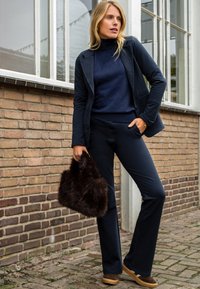 Marineblazer over een marineblauwe coltrui, gecombineerd met donkere flared broek. Houdt een donkerbruine nepbont handtas vast. Bruine schoenen met lichte zolen.