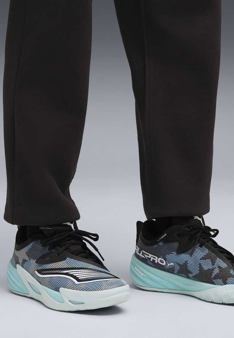 Personne portant un pantalon noir et des chaussures de sport noires et bleues à semelles bleu clair sur fond gris.