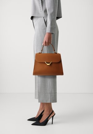Bolso de mano de cuero marrón con forma estructurada, herrajes dorados y asa superior. La modelo lleva un traje de cuadros y zapatos de tacón alto negros.