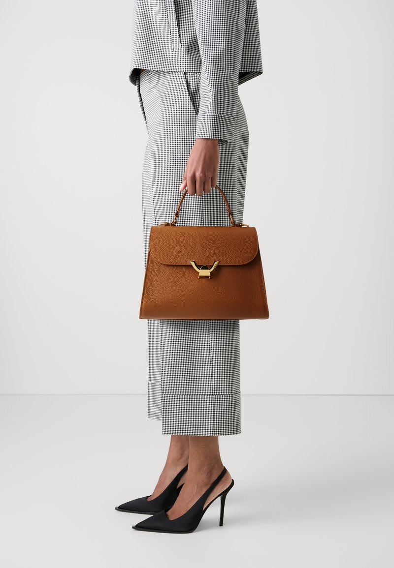 Borsa in pelle marrone con una forma strutturata, dettagli in oro e manico superiore. Il modello indossa un completo a quadri e scarpe con tacco nere.
