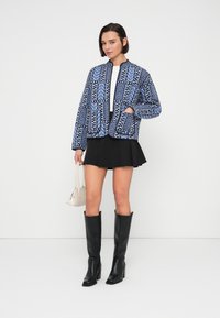 Μπλε και μαύρο μπουφάν μπόμπερ με φερμουάρ μπροστά, συνδυασμένο με μαύρη πλισέ φούστα και μαύρες δερμάτινες μπότες μέχρι το γόνατο. Κρατάει μια μικρή τσάντα.