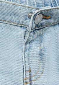 Blugi din denim albastru deschis în prim-plan, având un buton argintiu, cusături galbene contrastante și o textură netedă, cu o țesătură de material vizibilă.