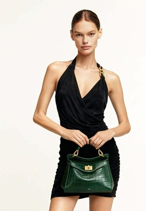 Femme en robe noire à licou tenant un sac à main vert à texture crocodile avec ferrures dorées, debout devant un fond clair uni.