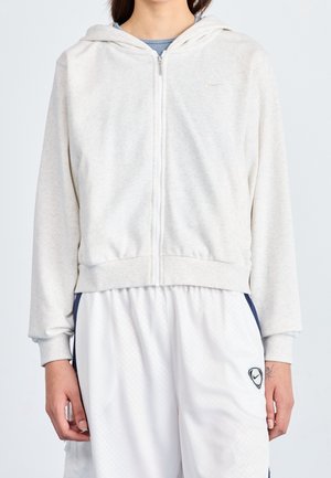 Person trägt einen hellgrauen Kapuzenpullover mit Reißverschluss und Kapuze, weiße Sportshorts mit marineblauen Seitenstreifen und Nike-Logo am linken Bein.