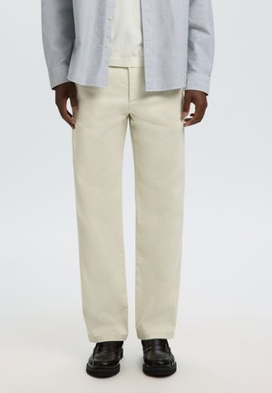 Homme portant un pantalon droit beige, des mocassins noirs, une chemise blanche et une chemise à rayures bleues et blanches boutonnée, debout devant un fond uni.