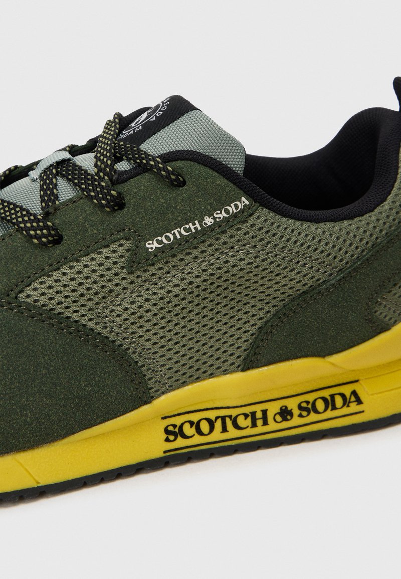 Grön sportig sneaker med mesh- och mockadetaljer, svarta accenter, gul sula och svarta snörningar med en texturerad yta och logodetalj.