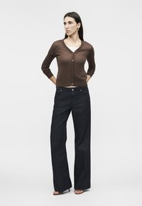 Γυναίκα όρθια με τα χέρια πίσω από την πλάτη, φοράει καφέ ζακέτα με κουμπιά, λευκό τοπ, τζιν με φαρδιά μπατζάκια και πέδιλα ανοιχτά στα δάχτυλα, μπροστά σε λευκό φόντο.