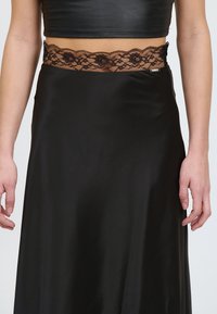 Gonna nera in satin con cintura in pizzo caratterizzata da motivi floreali e bordi ondulati. Tessuto morbido con un design fluido.