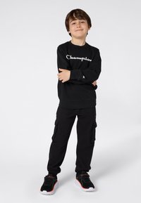 Chico de pie con confianza, con los brazos cruzados, vistiendo un sudadera negra de Champion, pantalones cargo negros y zapatillas negras con detalles en naranja.