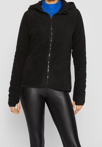 Zwarte fleecehoodie met een voorrits, zijzakken en een capuchon. Gecombineerd met glanzende zwarte leggings. Getextureerde stof, casual pasvorm.