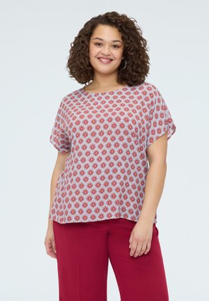 Uśmiechnięta kobieta o kręconych włosach, nosząca duże kolczyki-koła, bluzkę z krótkim rękawem w deseń oraz czerwone spodnie, stojąca na jednolitym tle.