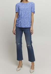 Top azzurro chiaro a maniche corte con un motivo floreale, abbinato a jeans blu con dettagli strappati. Modella che indossa scarpe con tacco beige.