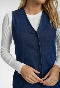 Tumedate teksast sinine denim vest, millel on V-kaeluse disain, ees nööbid ja kerge tekstuur. Koos valge pika varrukaga särgi all.