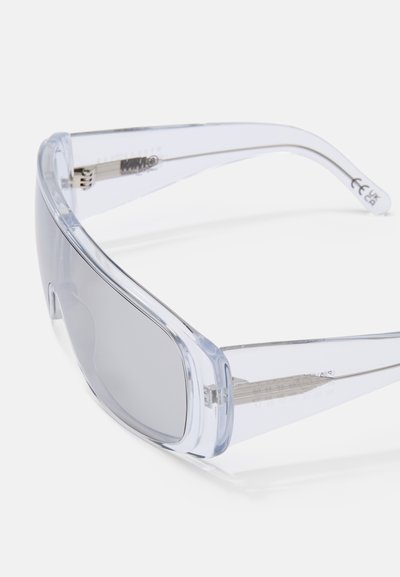Óculos de sol com uma estrutura de plástico transparente e leve e lentes cinza. Apresentam um design minimalista com detalhes discretos e bordas arredondadas.