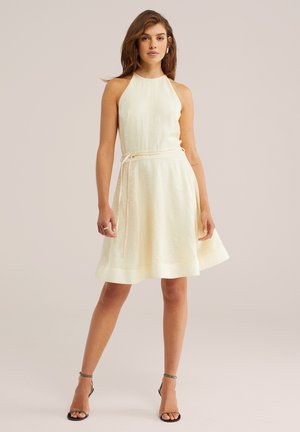 Kobieta w bezrękawowej, kremowej sukience o linii A z paskiem do wiązania, stojąca z rozstawionymi nogami, w czarnych butach na obcasie z paskiem wokół kostki, na neutralnym tle.