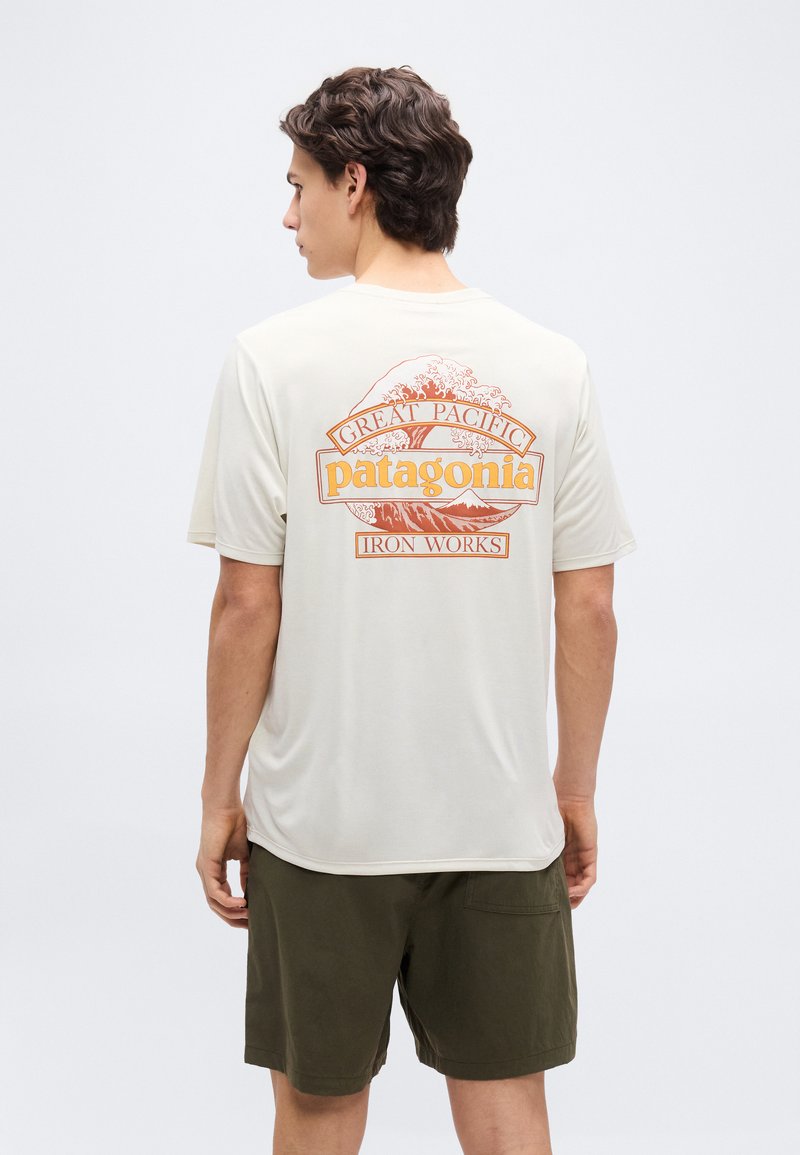 Νεαρός άνδρας που φοράει μπεζ μπλουζάκι Patagonia με πορτοκαλί κύμα και γραφικό βουνό, και χακί σορτς, στέκεται μπροστά σε απλό φόντο.